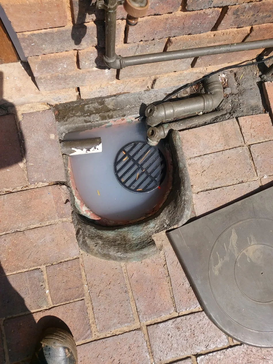 Outside gully drain filled with grey wastewater beside brick paving and pipework
