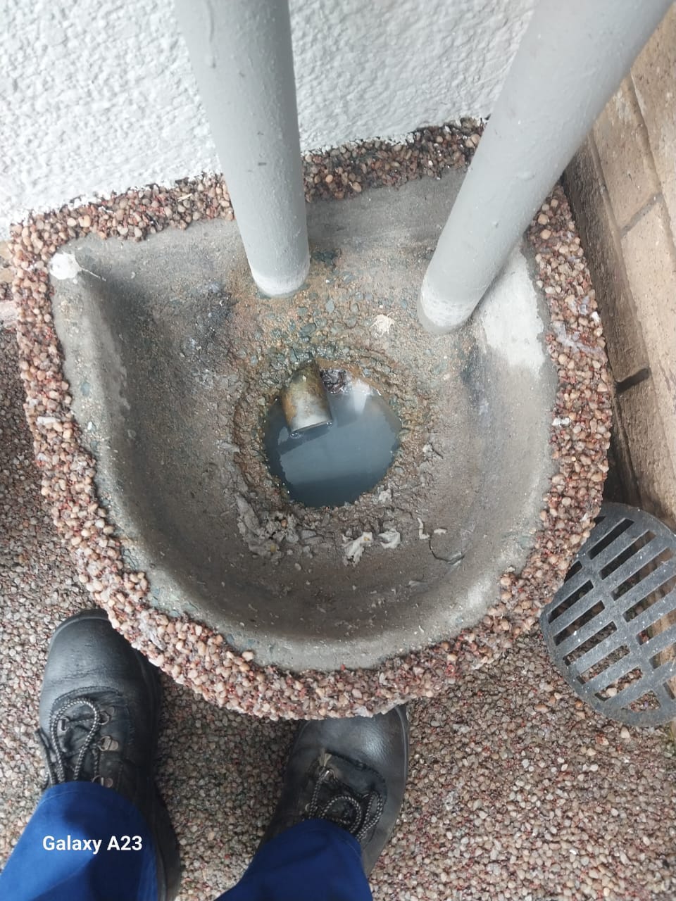 Blocked outdoor drain in Pomona with standing dirty water inside gully, surrounded by paving stones and downpipes, showing drainage blockage before cleaning and maintenance work.