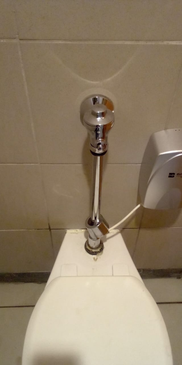 Chrome toilet flush valve installed on a commercial toilet with tiled wall background and wall mounted hand dryer, showing repaired flush mechanism and exposed flush pipe connection.