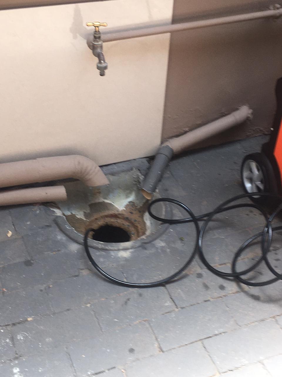 Blocked outdoor gulley drain beside wall-mounted tap with inspection cable inserted and surrounding pipes visible during drainage cleaning to restore proper water flow and prevent pooling.